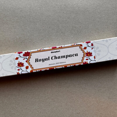 Räucherstäbchen-Verpackung 'Royal Champaca' von dhoop&art, Autumn 2024 Edition. Die längliche weiße Box zeigt ein elegantes Designelement in der Mitte - ein ornamentierter Rahmen mit dem Produktnamen, umgeben von dekorativen roten Blüten und orangefarbenen Blättern. Die Verpackung hat ein dezentes, helles Blumenmuster an beiden Enden.