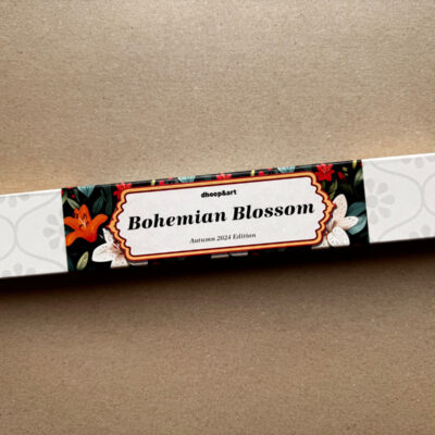 Räucherstäbchen-Verpackung 'Bohemian Blossom' von dhoop&art, Autumn 2024 Edition. Die längliche weiße Box zeigt ein elegantes Designelement in der Mitte - ein ornamentierter Rahmen mit dem Produktnamen auf schwarzem Hintergrund, umgeben von weißen Lilien und orangefarbenen Blüten mit grünen Blättern. Die Verpackung hat ein dezentes, helles Blumenmuster an beiden Enden.