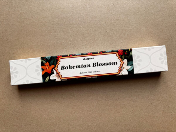 Räucherstäbchen-Verpackung 'Bohemian Blossom' von dhoop&art, Autumn 2024 Edition. Die längliche weiße Box zeigt ein elegantes Designelement in der Mitte - ein ornamentierter Rahmen mit dem Produktnamen auf schwarzem Hintergrund, umgeben von weißen Lilien und orangefarbenen Blüten mit grünen Blättern. Die Verpackung hat ein dezentes, helles Blumenmuster an beiden Enden.