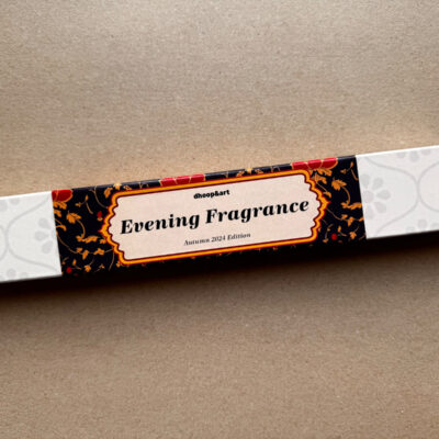 Räucherstäbchen-Verpackung 'Evening Fragrance' von dhoop&art, Autumn 2024 Edition. Die längliche weiße Box zeigt ein elegantes Designelement in der Mitte - ein ornamentierter Rahmen mit dem Produktnamen auf schwarzem Hintergrund, verziert mit roten Blüten und orangefarbenen Ranken. Die Verpackung hat ein dezentes, helles Blumenmuster an beiden Enden.