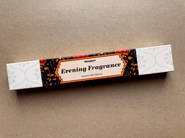 Räucherstäbchen-Verpackung 'Evening Fragrance' von dhoop&art, Autumn 2024 Edition. Die längliche weiße Box zeigt ein elegantes Designelement in der Mitte - ein ornamentierter Rahmen mit dem Produktnamen auf schwarzem Hintergrund, verziert mit roten Blüten und orangefarbenen Ranken. Die Verpackung hat ein dezentes, helles Blumenmuster an beiden Enden.