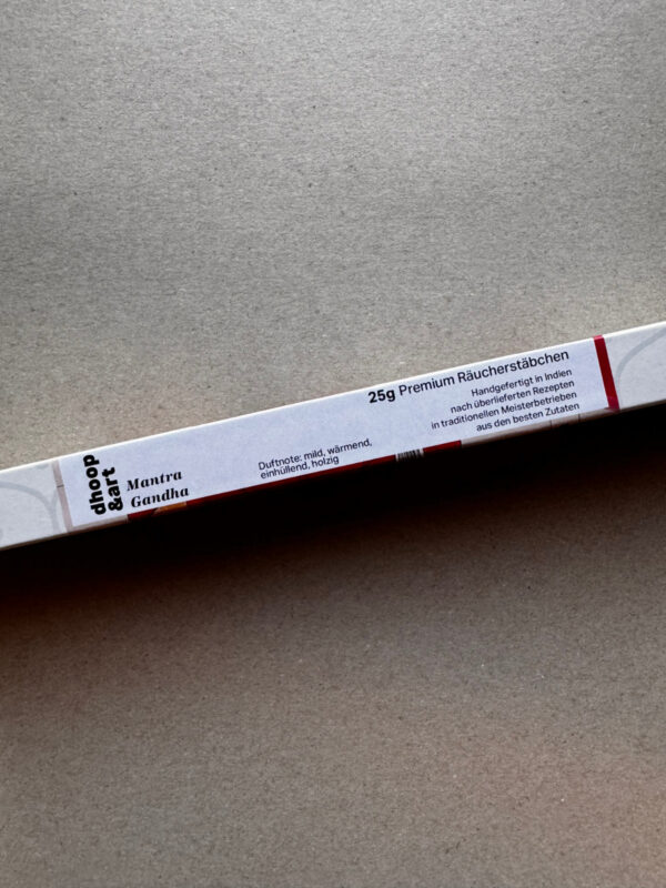 Detailansicht der dhoop&art 'Mantra Gandha' Räucherstäbchen-Verpackung. Zu sehen ist die Beschriftung mit Produktinformationen: '25g Premium Räucherstäbchen, handgefertigt in Indien nach überlieferten Rezepten in traditionellen Meisterbetrieben aus den besten Zutaten' sowie die Duftnote 'mild, wärmend, einhüllend, holzig'.